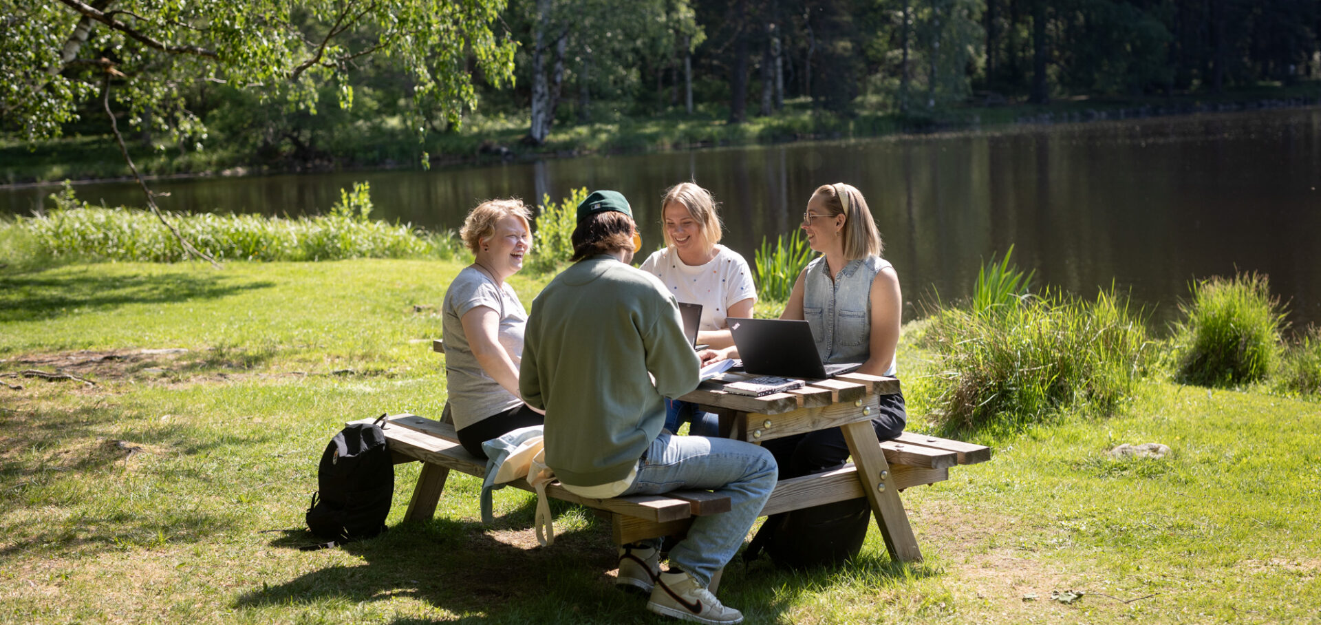 Neljä henkilöä istuu pöydän ääressä ja keskustelee lammen rannalla aurinkoisena kesäpäivänä.