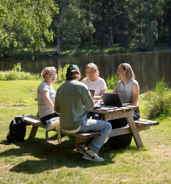 Neljä henkilöä istuu pöydän ääressä ja keskustelee lammen rannalla aurinkoisena kesäpäivänä.