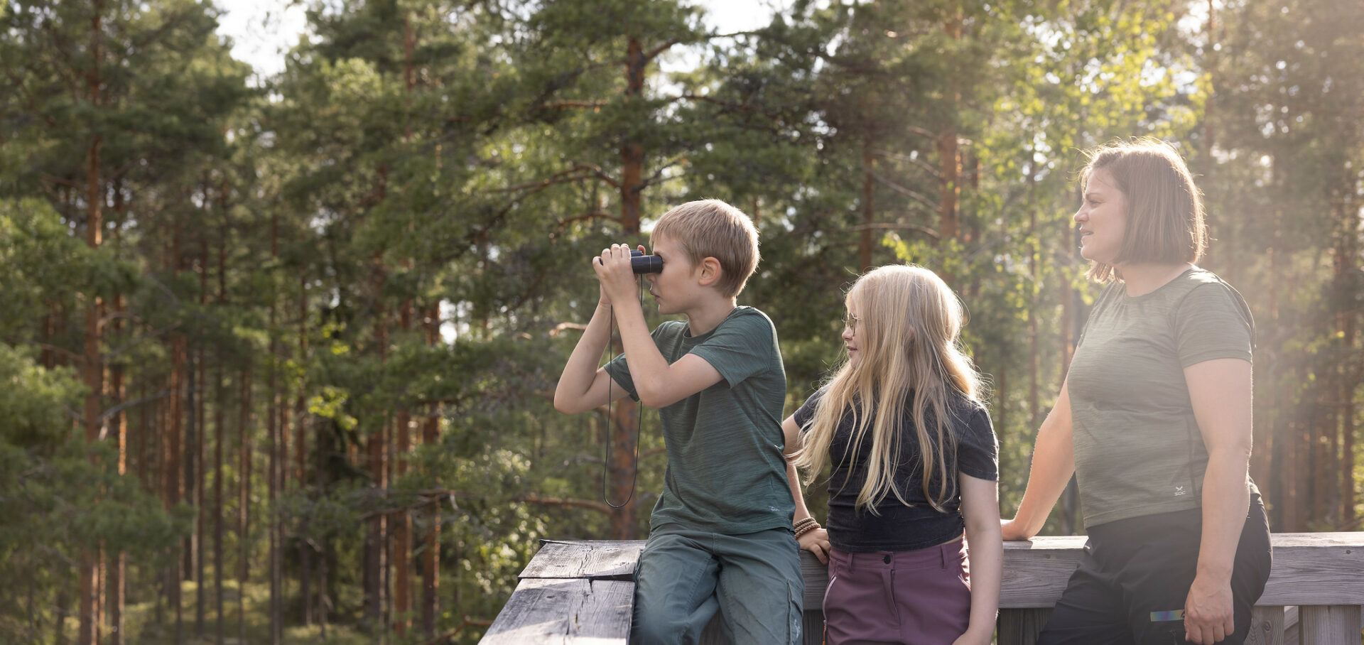 Edessä kaksi lasta, joista yksi kiikaroi, sekä aikuinen. Ihmisillä on päällä t-paidat ja housut. Taustalla kuusia.