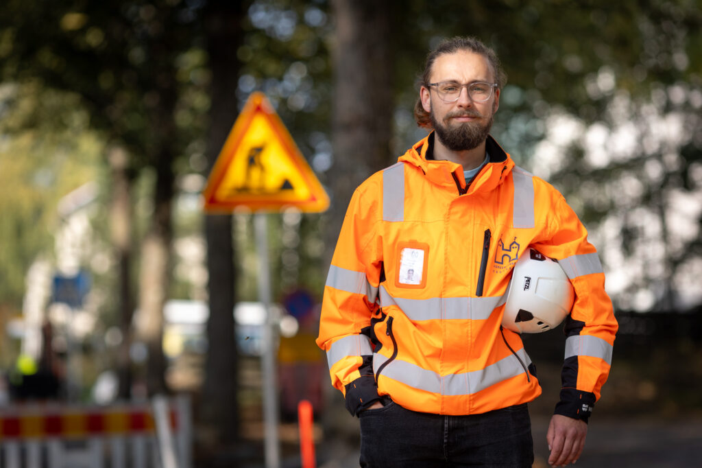 Hämeenlinnan kaupungin työntekijä seisoo työmaalla työmaatakki päällä ja kypärä kainalossa. Hänestä on otettu kuva uratarinaa varten.